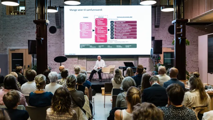 Norsk Fysioterapeutforbunds 90 års-markering. En sal med mange mennesker sittende på stoler. Lengst bak i bildet sitter en mann på en stol på en scene. Bak han er et stort projektorlerret hvor det vises frem noen fargede bokser med tekst.