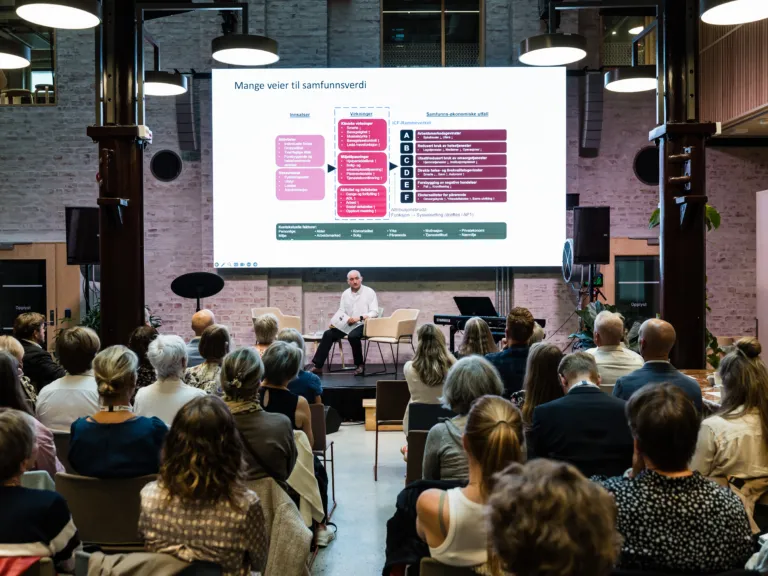 Norsk Fysioterapeutforbunds 90 års-markering. En sal med mange mennesker sittende på stoler. Lengst bak i bildet sitter en mann på en stol på en scene. Bak han er et stort projektorlerret hvor det vises frem noen fargede bokser med tekst.